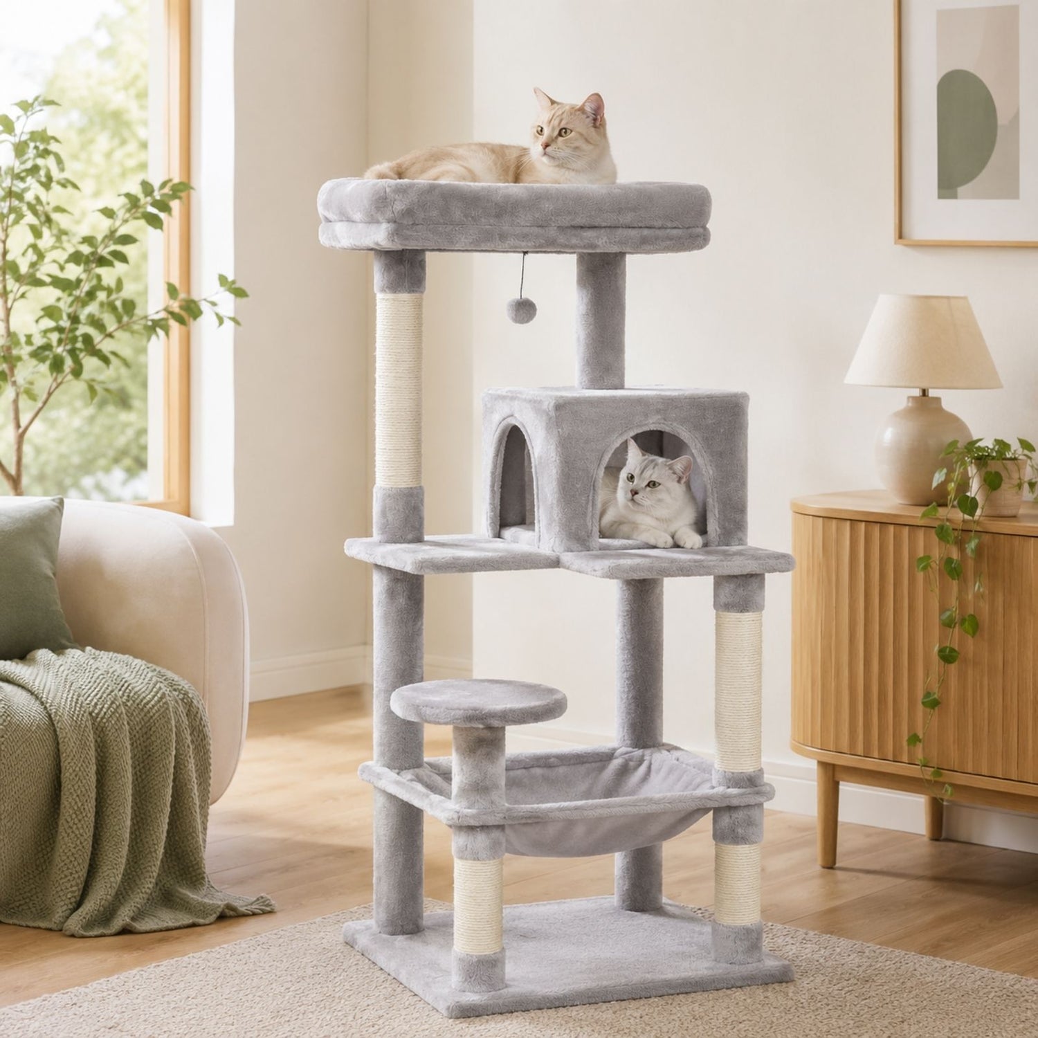A tall light gray cat tree with multiple levels, sisal-wrapped posts, a top platform with a cat lying on it, a small enclosed cat bed with a cat inside, a hanging toy, a lower platform, and a fabric hammock, shown in a bright living room with two cats.